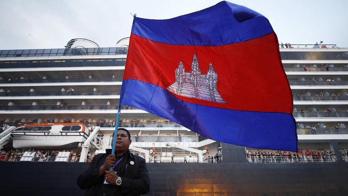 A Cambodian man holds his national flag in front of the MS Westerdam, owned by Holland America Line, dockked at the port of Sihanoukville, Cambodia, Friday, Feb. 14, 2020. Hundreds of cruise ship passengers long stranded at sea by virus fears cheered as they finally disembarked Friday and were welcomed to Cambodia. (AP Photo/Heng Sinith)