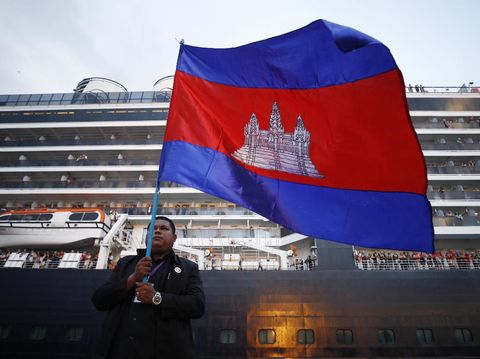 A Cambodian man holds his national flag in front of the MS Westerdam, owned by Holland America Line, dockked at the port of Sihanoukville, Cambodia, Friday, Feb. 14, 2020. Hundreds of cruise ship passengers long stranded at sea by virus fears cheered as they finally disembarked Friday and were welcomed to Cambodia. (AP Photo/Heng Sinith)