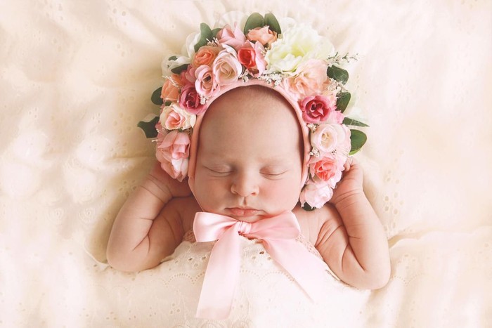 679510070 A newborn baby girl, fast asleep, wears a bonnet of flowers.