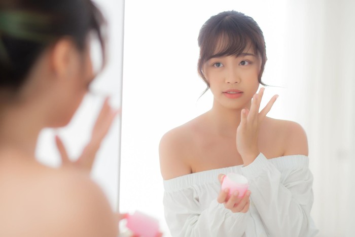 Beautiful portrait young asian woman applying cream moisturizer or lotion skin care cosmetic on face looking mirror, girl with treatment facial, health and wellness concept.