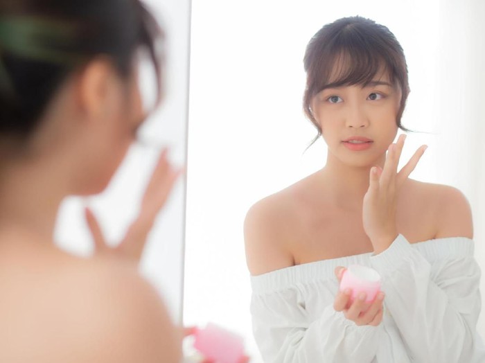 Beautiful portrait young asian woman applying cream moisturizer or lotion skin care cosmetic on face looking mirror, girl with treatment facial, health and wellness concept.