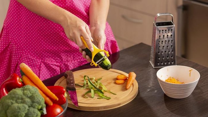 trik menggunakan peeler Julienned strands of a carrot are lying on a heart shaped chopping board. The Julienne utensil and a whole carrot are lying beside it on a rustic wooden surface. The peeler is made from stainless steel and has a black handle.