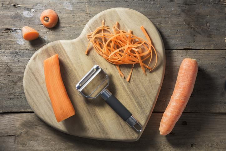 Julienned strands of a carrot are lying on a heart shaped chopping board. The Julienne utensil and a whole carrot are lying beside it on a rustic wooden surface. The peeler is made from stainless steel and has a black handle.