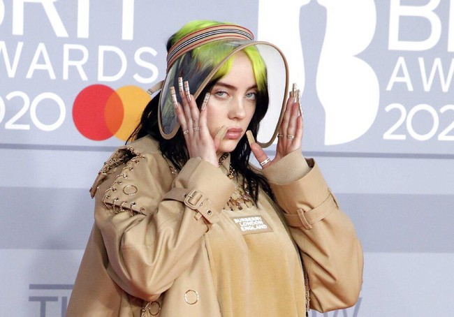 Untuk menyempurnakan gayanya, Billie memakai dad sneakers coklat berikut kaus kaki motif check khas Burberry. Tidak ketinggalan, topi transparan bergaya Amish yang menutup rambut hitam-hijau neonnya yang ekletik. (Foto: Vianney Le Caer/Invision/AP)