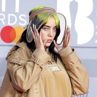 Untuk menyempurnakan gayanya, Billie memakai dad sneakers coklat berikut kaus kaki motif check khas Burberry. Tidak ketinggalan, topi transparan bergaya Amish yang menutup rambut hitam-hijau neonnya yang ekletik. (Foto: Vianney Le Caer/Invision/AP)