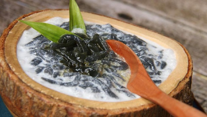 Es Dawet Ireng, the Javanese cold dessert of black cendol (tapioca short-noodle jelly) made from toasted paddy straw. It is served with crushed ice, coconut milk, and palm sugar syrup in a thick wooden bowl that has been carved out of single wooden block. A wooden spoon is in the midst of stirring to mix the palm sugar syrup and and coconut milk. The dessert is arranged on a rustic wooden table.