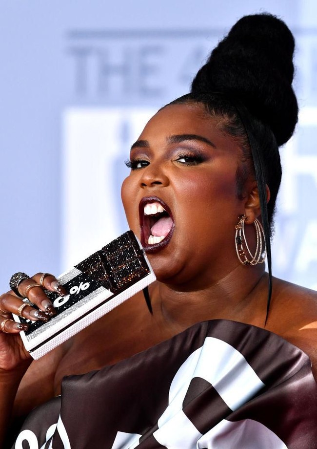 Lizzo lalu menyempurnakan gayanya dengan clutch berbentuk batangan coklat dengan tulisan 100%. Besar kemungkinan angka tersebut diambil dari lirik lagunya yang berjudul Truth Hurts: Im 100 percent that b----. (Foto: Gareth Cattermole/Getty Images)