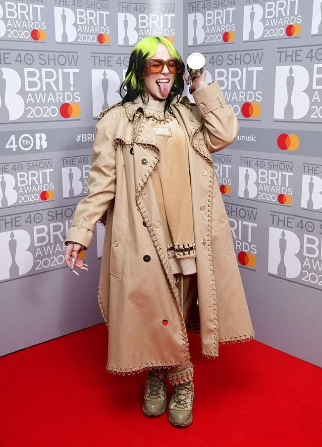 Billie Eilish membungkus tubuhnya dengan tracksuit coklat yang dihiasi dengan detail cincin yang berjajar di area neckline dan keliman atasannya. Busana rancangan Riccardo Tisci itu lalu dilengkapi luaran berupa trench coat klasik Burberry dalam versi modern dengan detail cincin yang sama. (Foto: Ian West/ PA via AP)