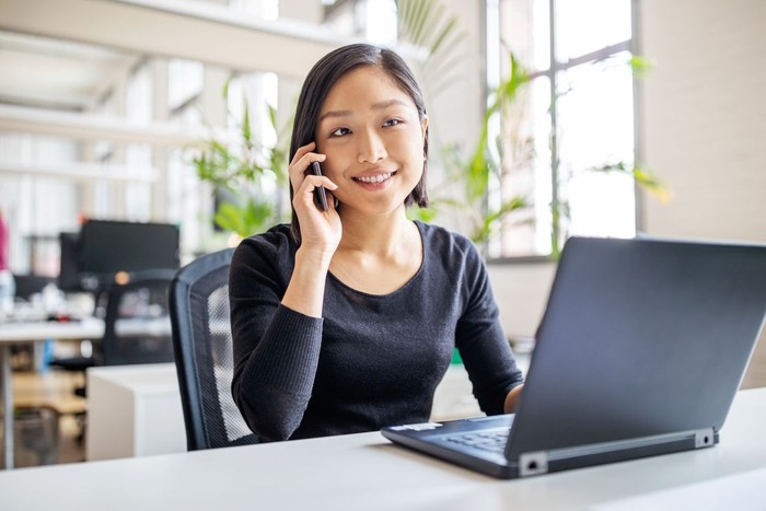 Young asian businesswoman sitting at her desk with laptop talking on mobile phone. Female professional working in modern office.