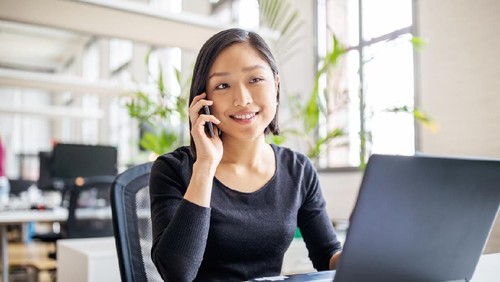 Young asian businesswoman sitting at her desk with laptop talking on mobile phone. Female professional working in modern office.