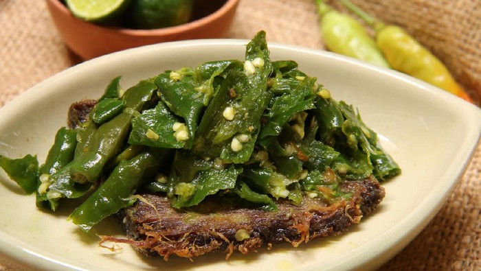 A popular Padangnese dish of dried beef jerky topped with green chili. Plated on a oval ceramic plate. In the background are a bowl of limes and chili peppers.