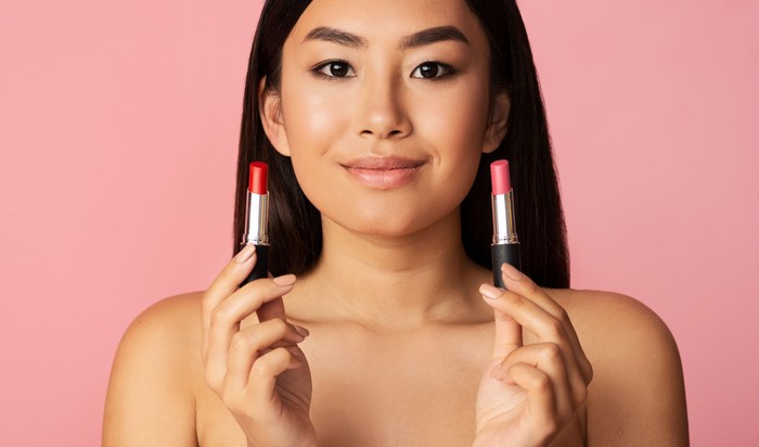 lipstik untuk kulit sawo matang Makeup concept. Asian girl holding two lipsticks over pink studio background, panorama