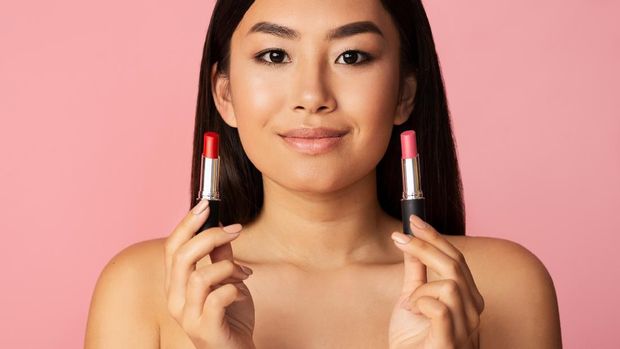 Makeup concept. Asian girl holding two lipsticks over pink studio background, panorama