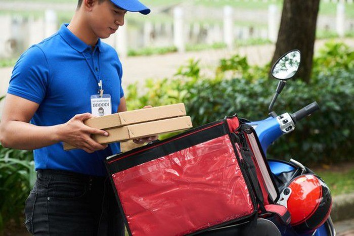 Young male courier in yellow sweatshirt delivering pizza, standing with thermal backpack and smart phone on the steret outdoors