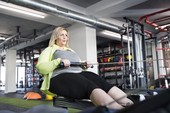 682197326 Attractive overweight woman in modern gym exercising on rowing machine. Chubby young blonde woman at fitness center.