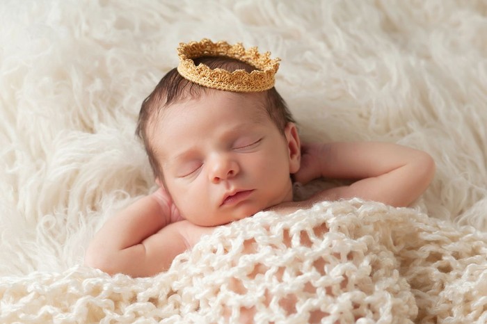 474094423 Portrait of a 12 day old newborn baby boy wearing a gold crown. He is sleeping on a beige flokati rug with his hands behind his head.