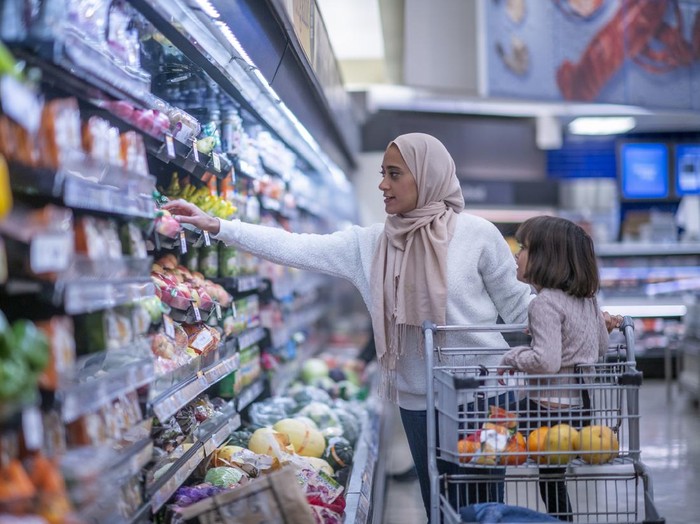 A young Muslim mother and daughter walk the produce aisles of the grocery store together shopping for groceries.  They are both dressed casually and the mother is wearing a Hijab.  The mother is reaching for products on the shelf while pushing a shopping cart with her daughter seated in it.