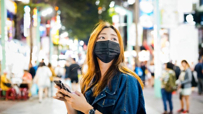 Woman wearing pollution mask while using mobile phone. Young female is looking away while standing on street. She is in city at night.