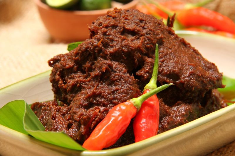 Rendang Padang, the most famous Padangnese dish of spicy dry beef stew. Plated on a rectangular ceramic plate that has been lined with banana leaf. Two chili peppers are used to garnish the dish.