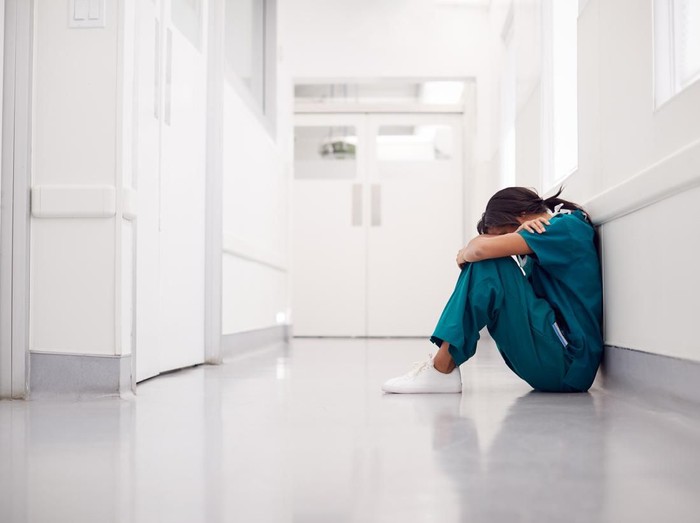 Stressed And Overworked Female Doctor Wearing Scrubs Sitting On Floor In Hospital Corridor