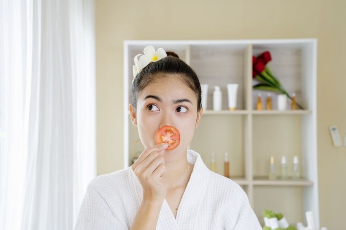 masker wajah Young beautiful wearing a white bathrobes in a relaxed girl smiling hiding eye behind tomato slice over spa room background. Beauty spa and cosmetology concept. casual atmosphere.