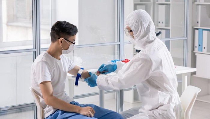 Young Chinese man in mask sitting in lab while doctor in protective coveralls taking his blood in syringe for analysis