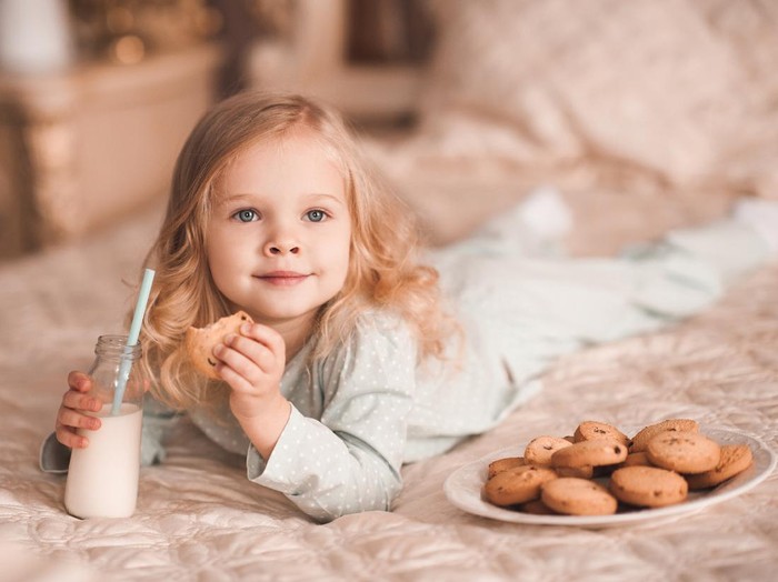 Sweet baby girl 3-4 year old eating cookies and drinking milk lying in bed close up. Good morning. Breakfast.