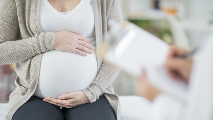 Ibu hamil A pregnant Caucasian woman is indoors in a doctors office. Her female doctor is wearing medical clothing. The woman is holding her stomach which the doctor writes on a clipboard.
