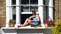Seorang pria menikmati waktunya di rumah dengan berjemur sembari membaca dan mendengarkan musik dair jendela rumahnya. Foto ini diambil di London, 5 April 2020. (REUTERS/Dylan Martinez)