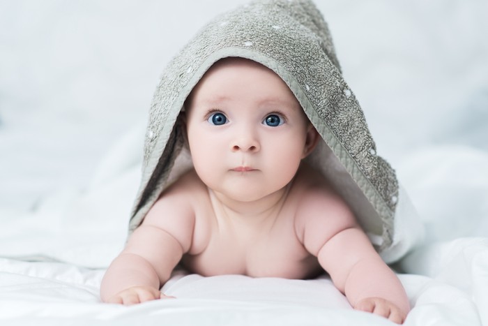 Cute baby girl or boy after shower with towel on head in white sunny bedroom. Child with big blue eyes relaxing in bed after bath