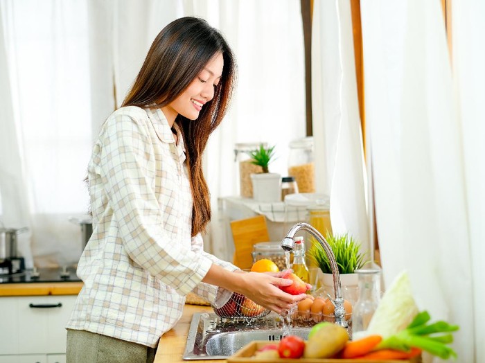 Cara mencuci bahan makanan yang tepat Asian beautiful teen girl is washing the apple and other fruit in the kitchen and look happy.