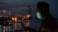 Warga dengan mengenakan masker tampak dalam frame dengan latar belakang Jembatan Suramadu yang lampunya sudah mulai menyala.