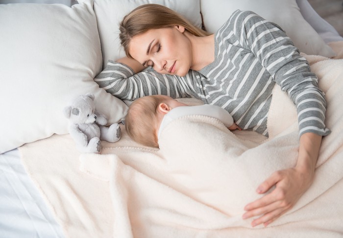 Beautiful mom lying at the back of the baby while the infant is sleeping on the white bed. Asian mother looking at the baby which is her child with love and care.