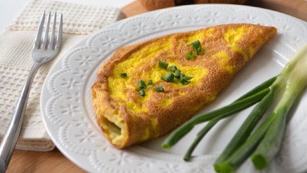 Fluffy breakfast omelette with green spring onions and fresh toast bread on a wooden surface with a napkin and metal fork