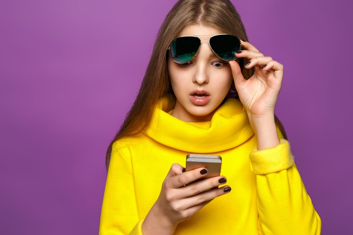 Portrait surprised young girl from message on smartphone in brightly yellow sweater, isolate on a violet background