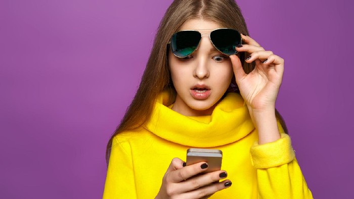 Portrait surprised young girl from message on smartphone in brightly yellow sweater, isolate on a violet background