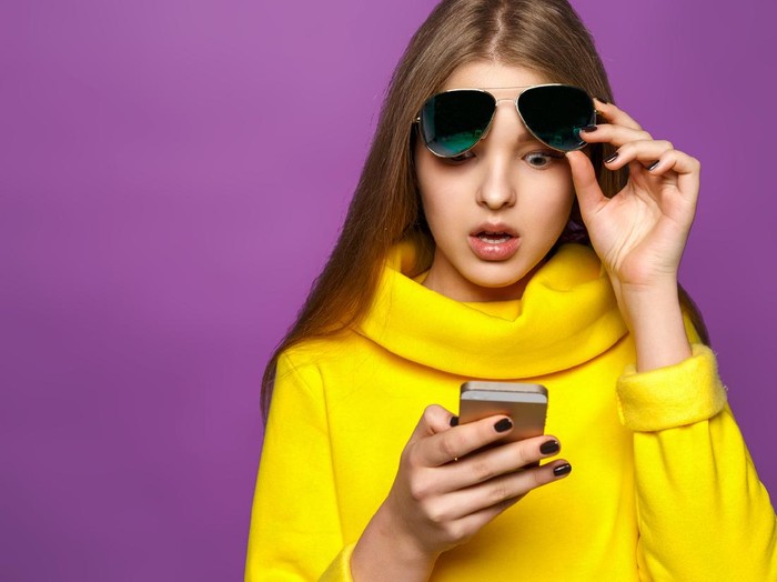 Portrait surprised young girl from message on smartphone in brightly yellow sweater, isolate on a violet background