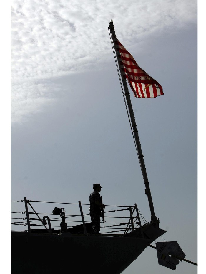 FILE- In this Jan. 12, 2012 file photo, a U.S. Navy sailor is seen by the American flag during a change-of-command ceremony Taboard the destroyer USS Kidd docked in Muharraq, Bahrain. The Navy says another warship at sea has reported a coronavirus outbreak and is returning to port. In a written statement, the Navy said Friday at least 17 members of the crew of the USS Kidd have tested positive for the virus. The destroyer was operating in the Caribbean Sea.  (AP Photo/Hasan Jamali)
