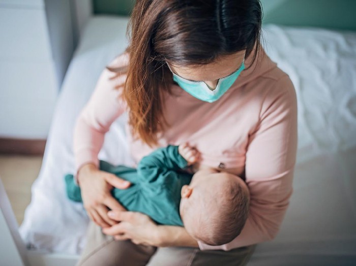 Two people, mother with protective mask breastfeeding her baby son at home, they are at home do to pandemic outbreak quarantine.