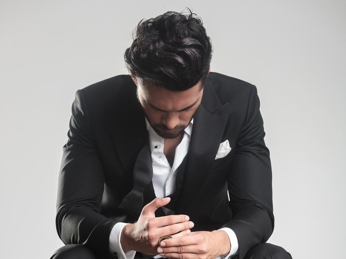 Pria Ditipu Calon Istri Elegant young man in tuxedo looking down while sitting on a stool, on grey studio background.