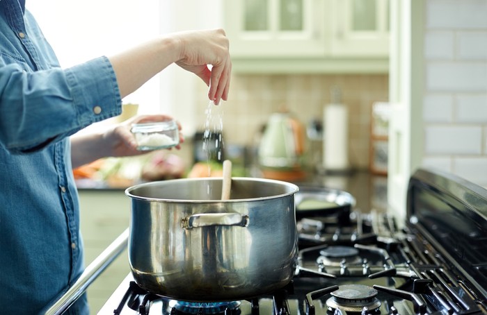 Midsection image of woman adding salt in cooking pot. Utensil is placed on gas stove. Close-up of female seasoning her dish. She is preparing food in domestic kitchen.