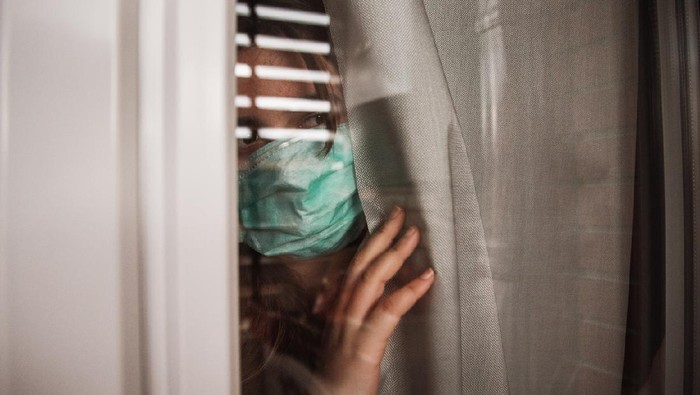 A young woman in quarantine wearing a mask and looking through the window
