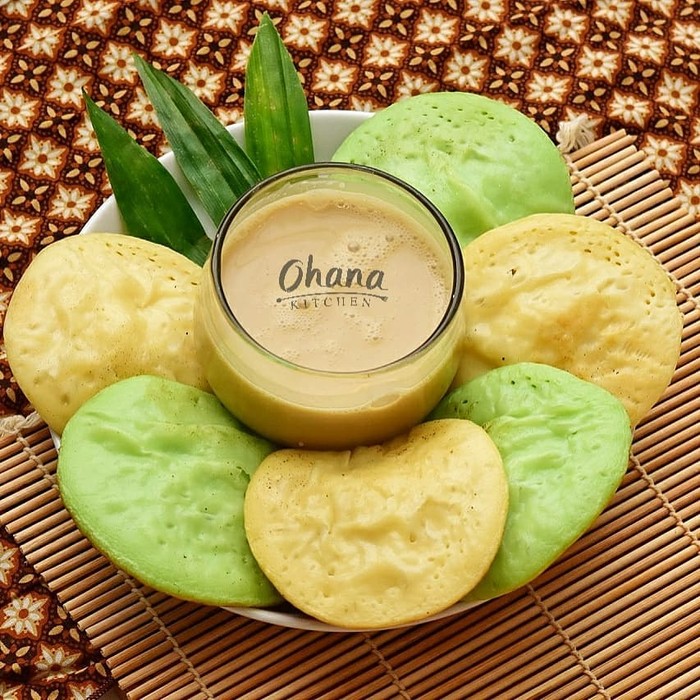 Javanese coconut milk and pandanus pancakes, served with sauce of coconut milk and palm sugar. Both the pancakes and the sauce are plated on traditional earthenware crockery set. Two wooden spoon are placed next to the plate. A folded woven straw mat lines the plate and cup. The meal is arranged on rustic wooden table that has a matching color with the earthenware. The image was taken outdoor using natural light.