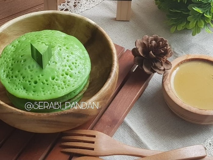 Javanese coconut milk and pandanus pancakes, served with sauce of coconut milk and palm sugar. Both the pancakes and the sauce are plated on traditional earthenware crockery set. Two wooden spoon are placed next to the plate. A folded woven straw mat lines the plate and cup. The meal is arranged on rustic wooden table that has a matching color with the earthenware. The image was taken outdoor using natural light.