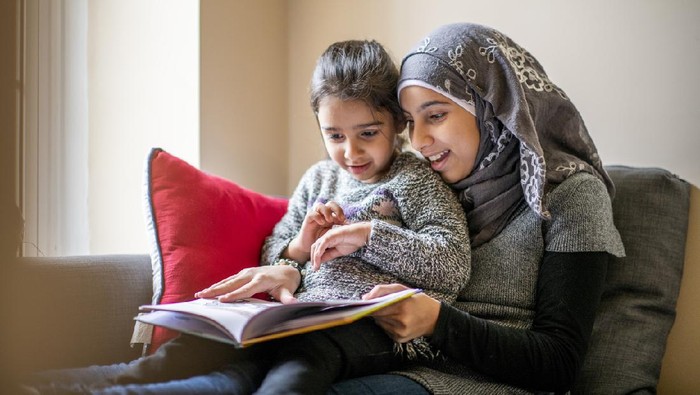 A pre-teen girl wearing a hijab sits on a couch with her little sister on her lap and reads her a bedtime story. Her sister is engaged in the story.