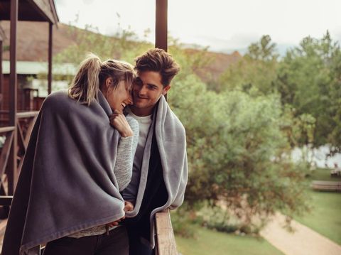 1012469644 Loving man and woman standing in their hotel room balcony wrapped in blanket. Romantic couple in a blanket standing together on a winter holiday.