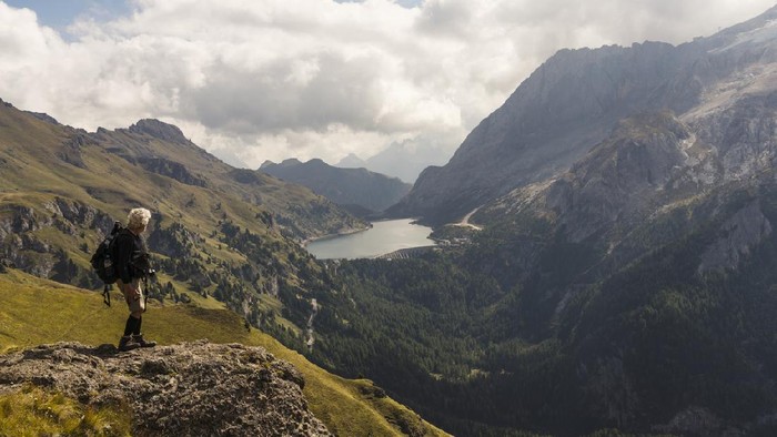 Senior man hiking along hills opposite to the Marmolada in autumn. In distance lake Fedaia. Dolomites, Northern Italy.