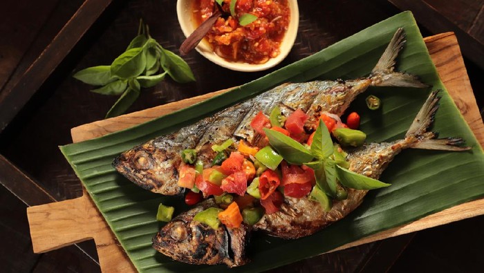 Ikan Tude Bakar Sambal Dabu, a popular Minahasan dish of char-grilled mackerel topped with Dabu-dabu salsa, which is the Minahasan fresh salsa of tomatoes, chili peppers, and shallot. The fish is plated on rectangular wooden board lined with banana leaf. A small bowl of Sambal Rica (Minahasan red chili pepper paste) is served as extra condiment to the grilled fish.