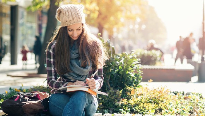 Student girl writing in notebook and studying. Casual clothing, scarf and hat.