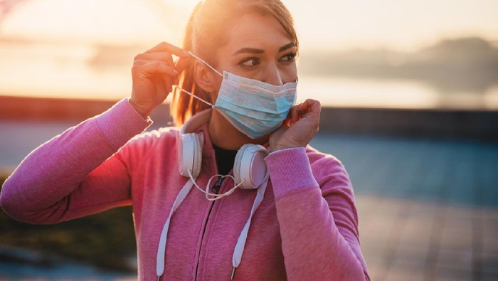 joging jogging lari pakai masker Beautiful female jogger putting protective mask on her face to protect herself from virus or allergy infection. Sunset in background.